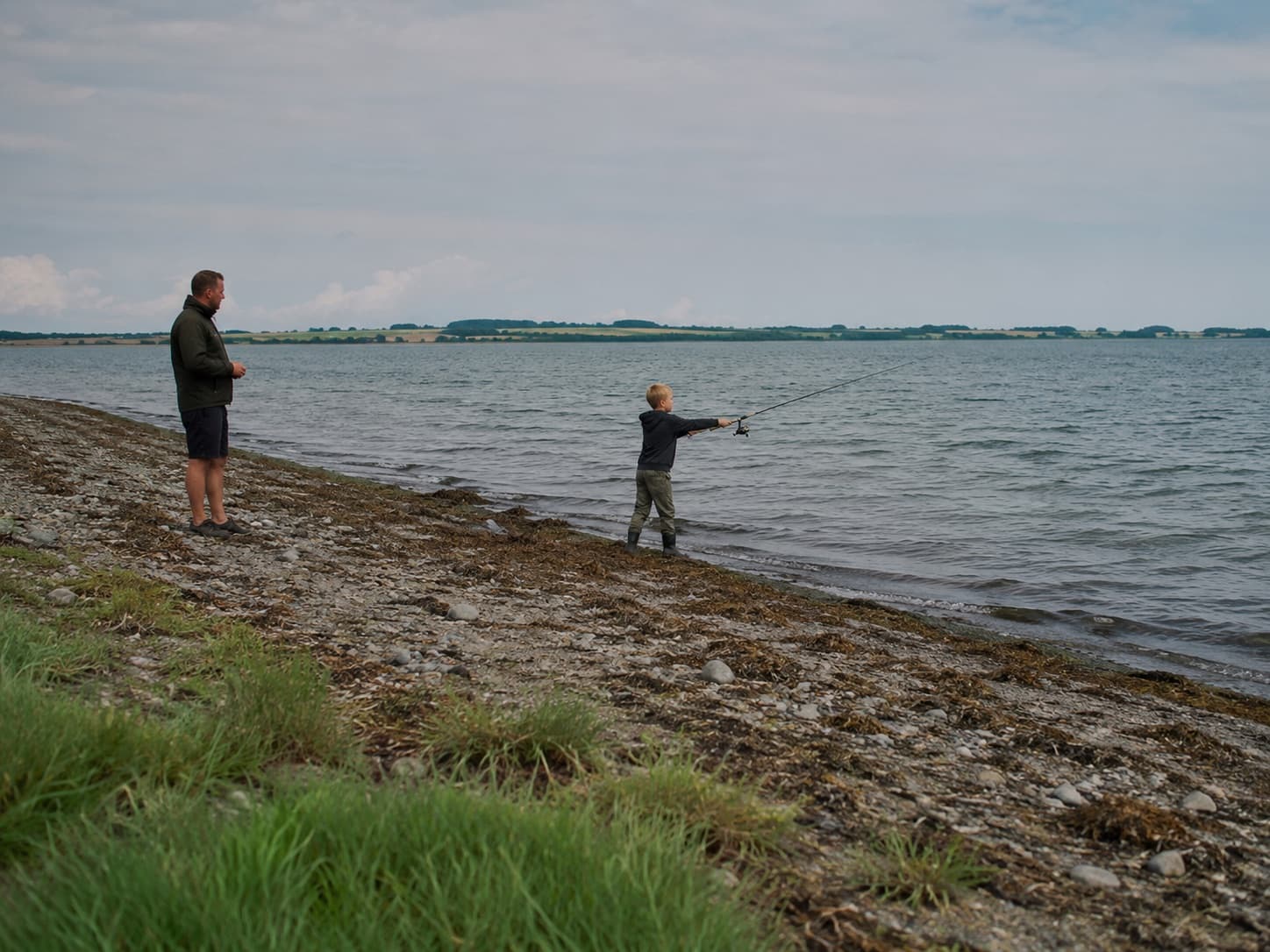 Dreng kaster selv med spinnestang fra en bred strand, mens en voksen holder afstand og kigger på