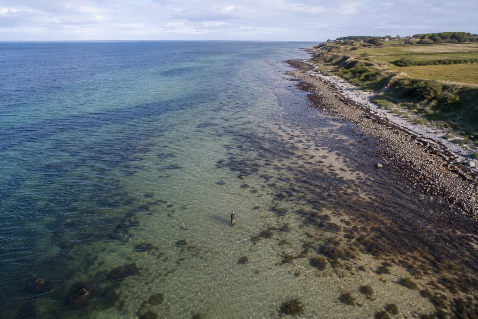 Luftfoto af en lystfisker, der vader i krystalklart vand langs en stenet kyststrækning