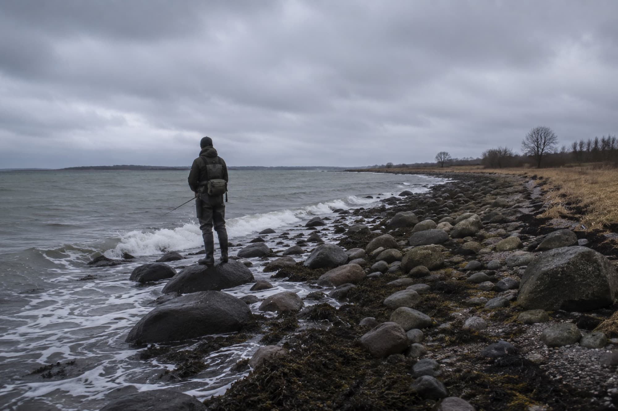 Lystfisker står på en sten ved en overskyet, stenet kyst med bølger, der bryder