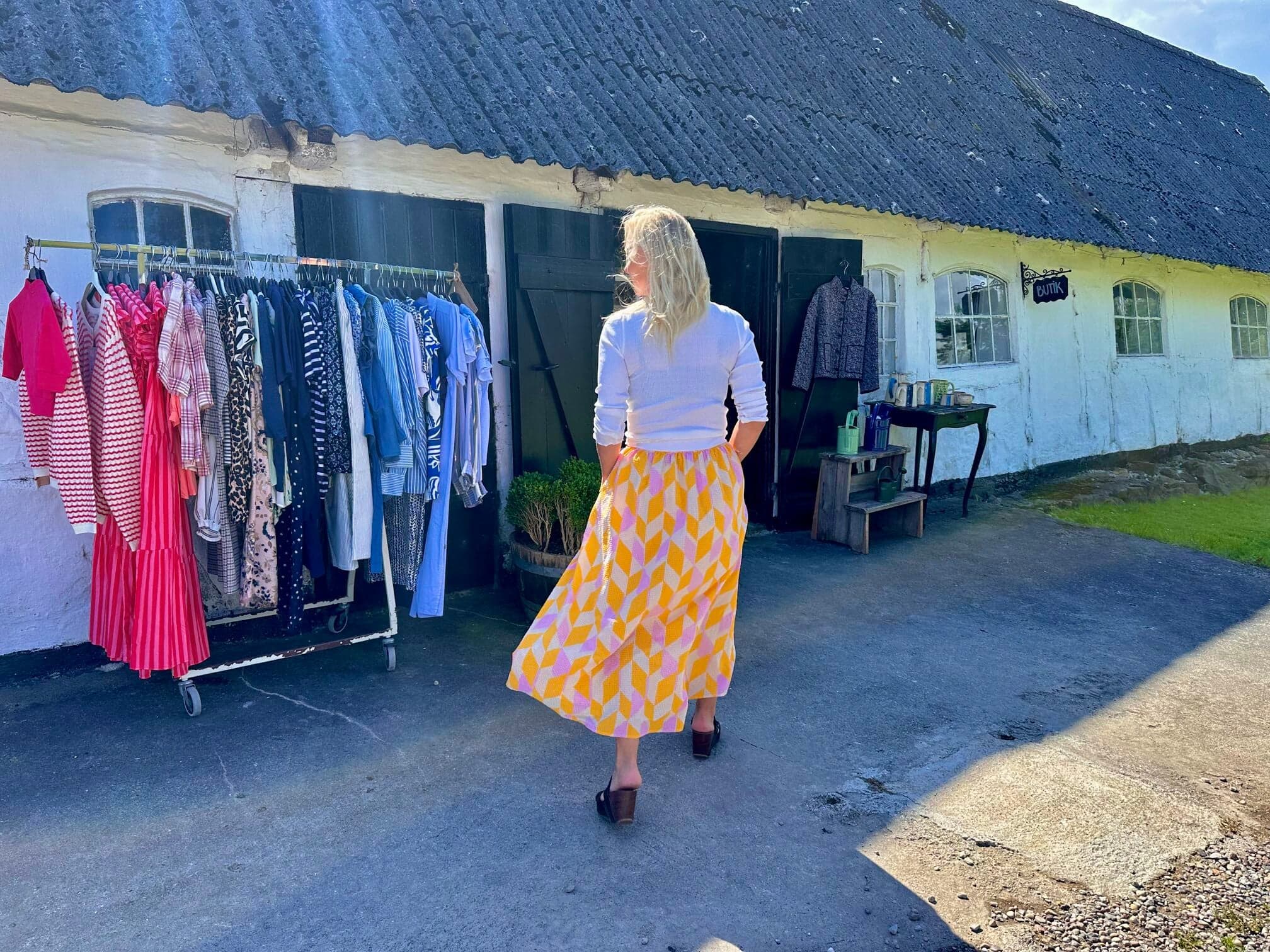 En gæst går ind i butikken hos Himmel & Hav, med tøjstativer og skiltet Butik på den hvide bindingsværksbygning